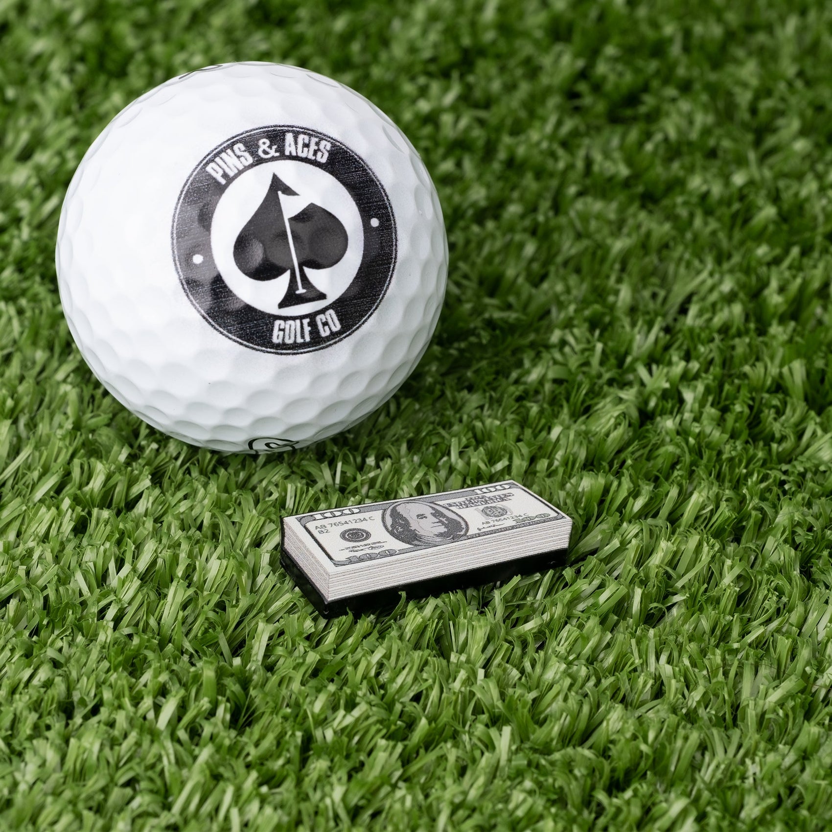 A white golf ball with a Pins and Aces logo sits on artificial grass next to novelty $100 bills and the Cash Stack Ball Marker from Pins and Aces.