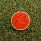 The Logie Joe's Golf Watermelon "Slice" Premium Ball Marker features a round, watermelon slice design with a removable segment, displayed on green artificial grass.