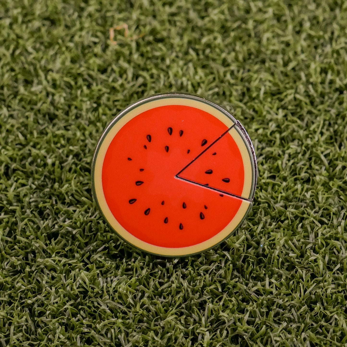 The Logie Joe's Golf Watermelon "Slice" Premium Ball Marker features a round, watermelon slice design with a removable segment, displayed on green artificial grass.