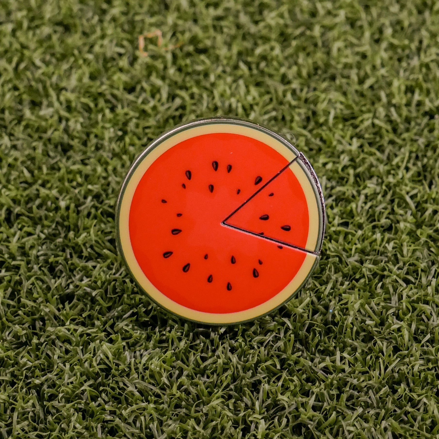 The Logie Joe's Golf Watermelon "Slice" Premium Ball Marker features a round, watermelon slice design with a removable segment, displayed on green artificial grass.