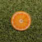 The Logie Joe's Golf Orange "Slice" Premium Ball Marker, shaped like an orange slice with a removable piece, sits on textured green grass.