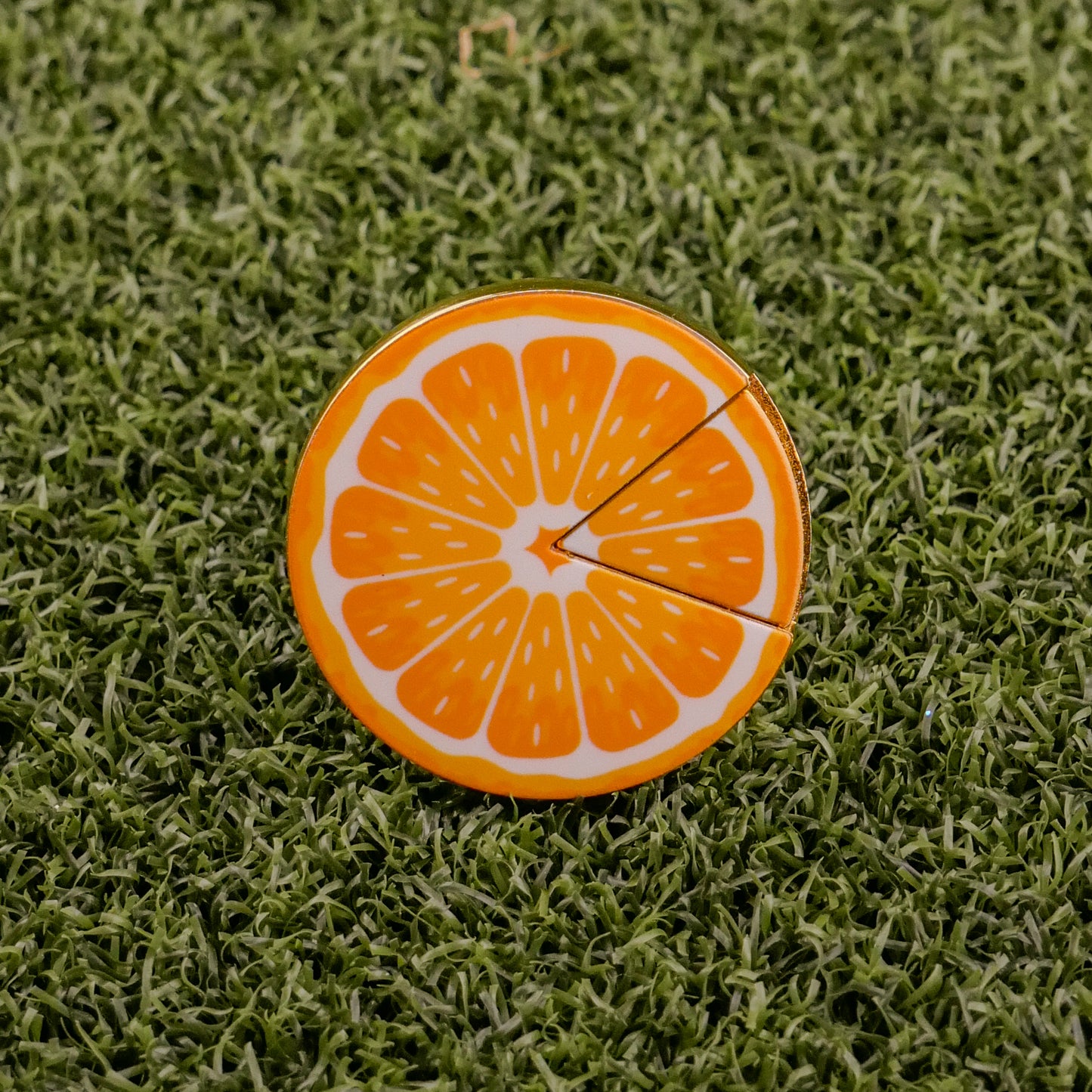 The Logie Joe's Golf Orange "Slice" Premium Ball Marker, shaped like an orange slice with a removable piece, sits on textured green grass.