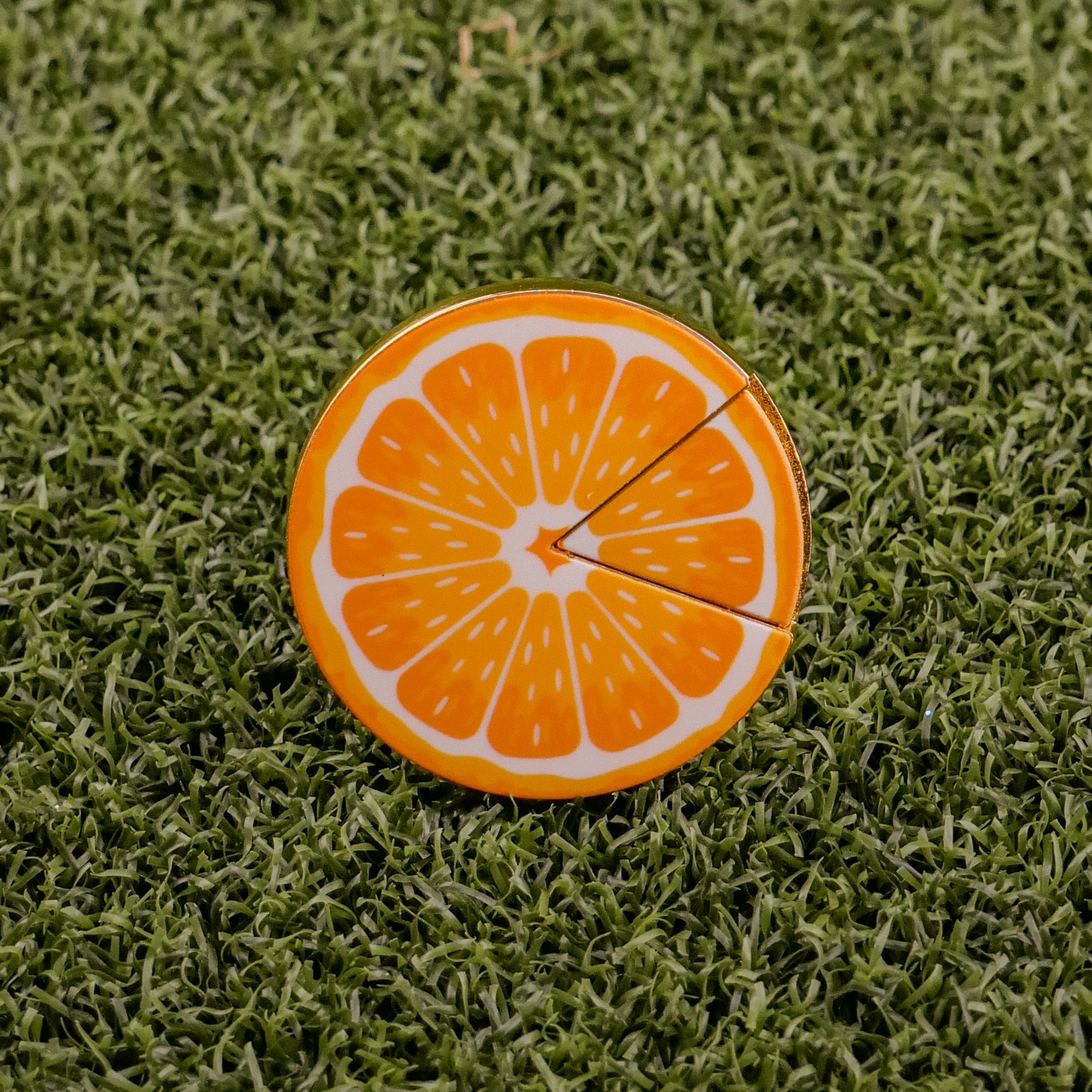 The Logie Joe's Golf Orange "Slice" Premium Ball Marker, shaped like an orange slice with a removable piece, sits on textured green grass.