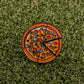 The Supreme Pizza "Slice" Premium Ball Marker by Logie Joe's Golf is a round enamel pin featuring pizza toppings and a removable slice, ideal as a quirky golf or poker chip ball marker on green grass surfaces.