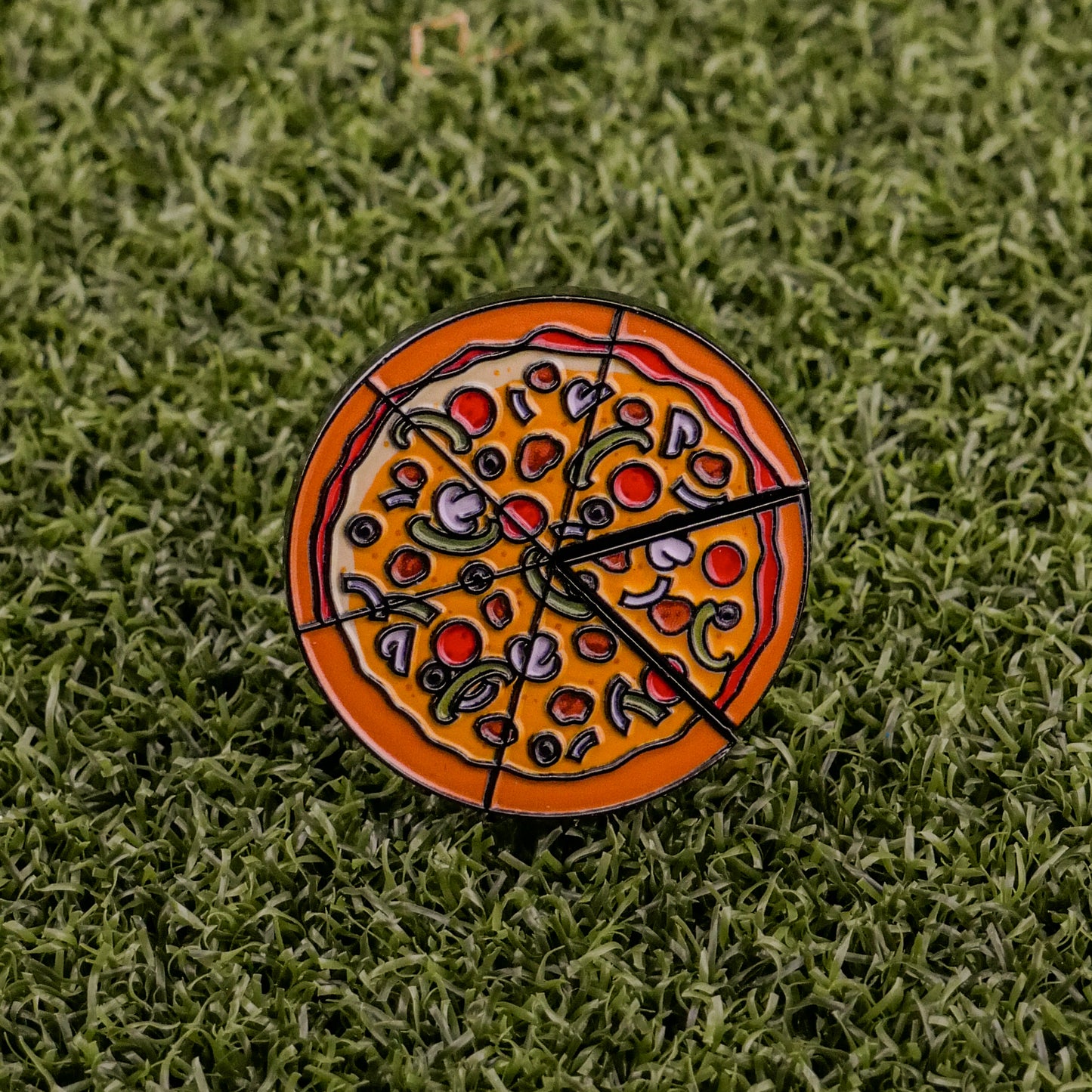 The Supreme Pizza "Slice" Premium Ball Marker by Logie Joe's Golf is a round enamel pin featuring pizza toppings and a removable slice, ideal as a quirky golf or poker chip ball marker on green grass surfaces.