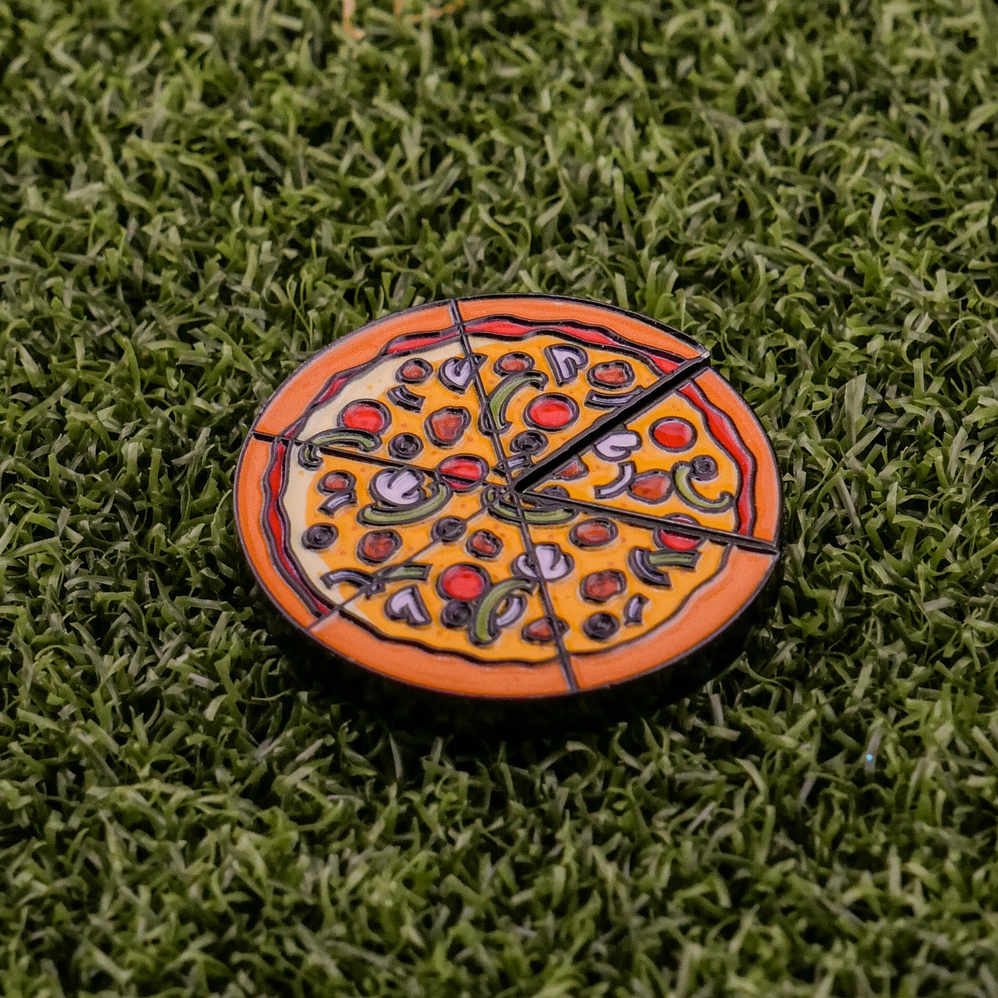 The Logie Joe's Golf Supreme Pizza "Slice" Premium Ball Marker, featuring black slice markings and removable slice detail, rests on artificial green grass.
