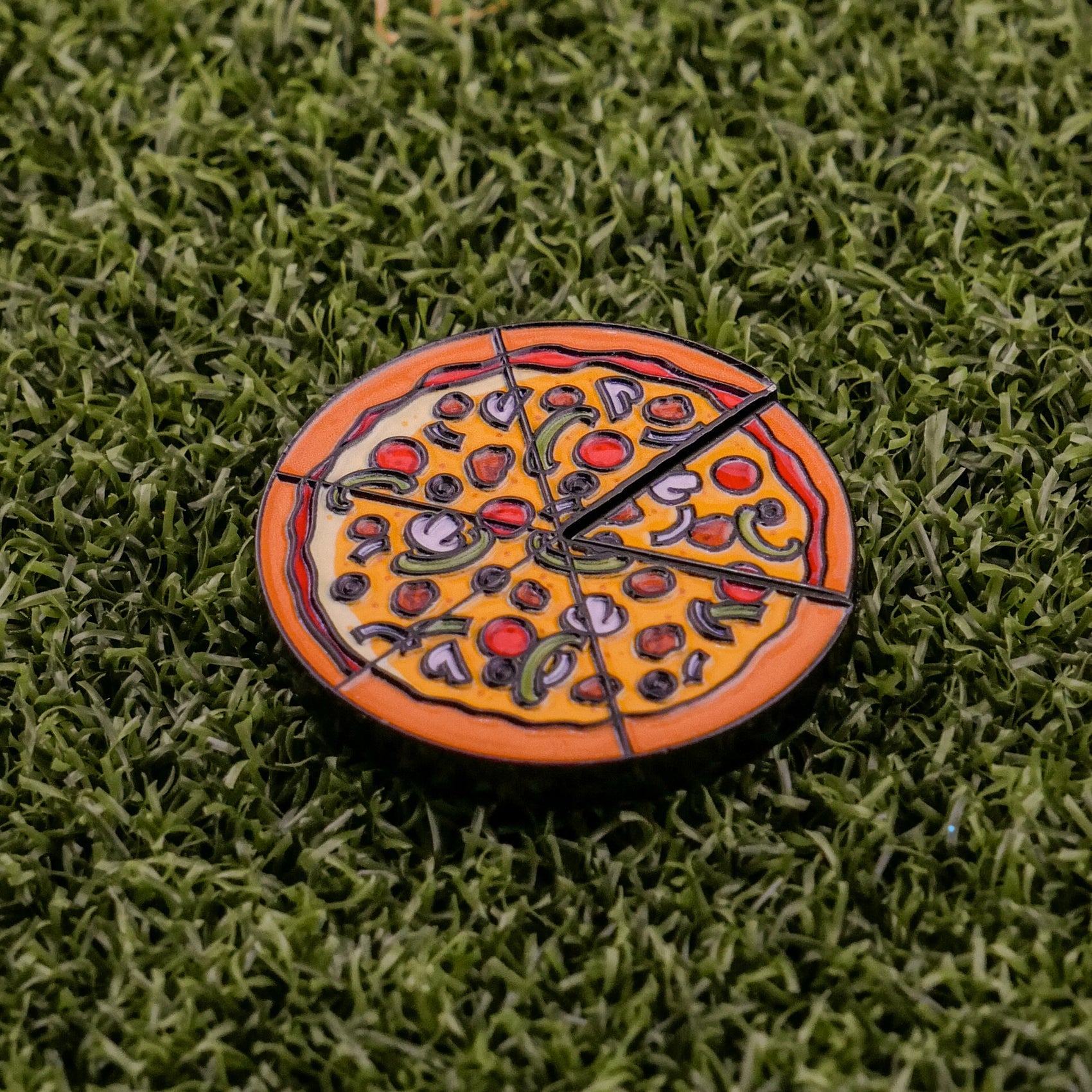 The Logie Joe's Golf Supreme Pizza "Slice" Premium Ball Marker, featuring black slice markings and removable slice detail, rests on artificial green grass.