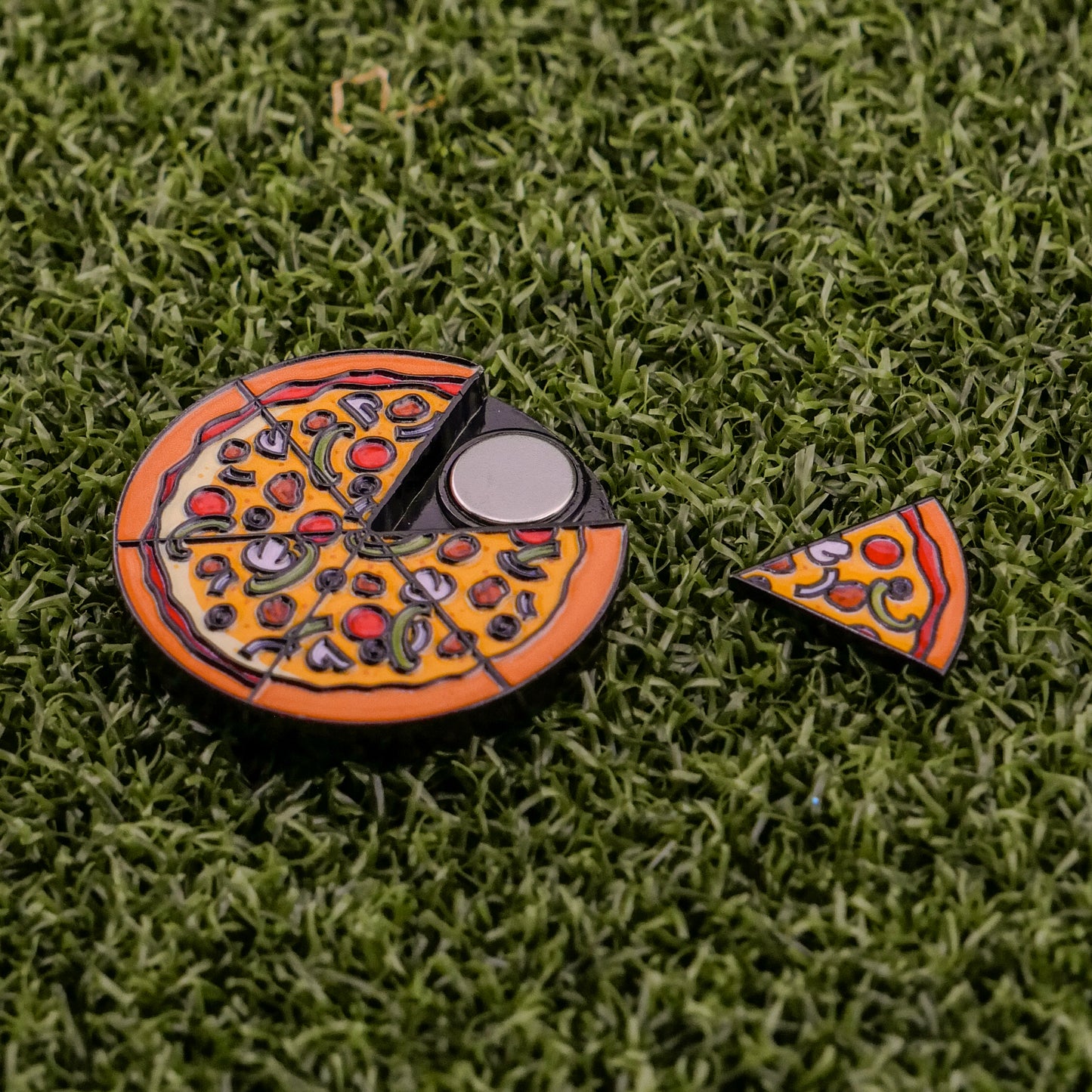 The Logie Joe's Golf Supreme Pizza "Slice" Premium Ball Marker, shaped like a pizza with colorful toppings and a removable slice, sits on green artificial grass, showing a black circular section where the slice was removed.