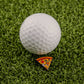 A white golf ball sits on green turf beside the Logie Joe's Golf Supreme Pizza "Slice" Premium Ball Marker, designed as a small triangular pizza slice with pepperoni and cheese.