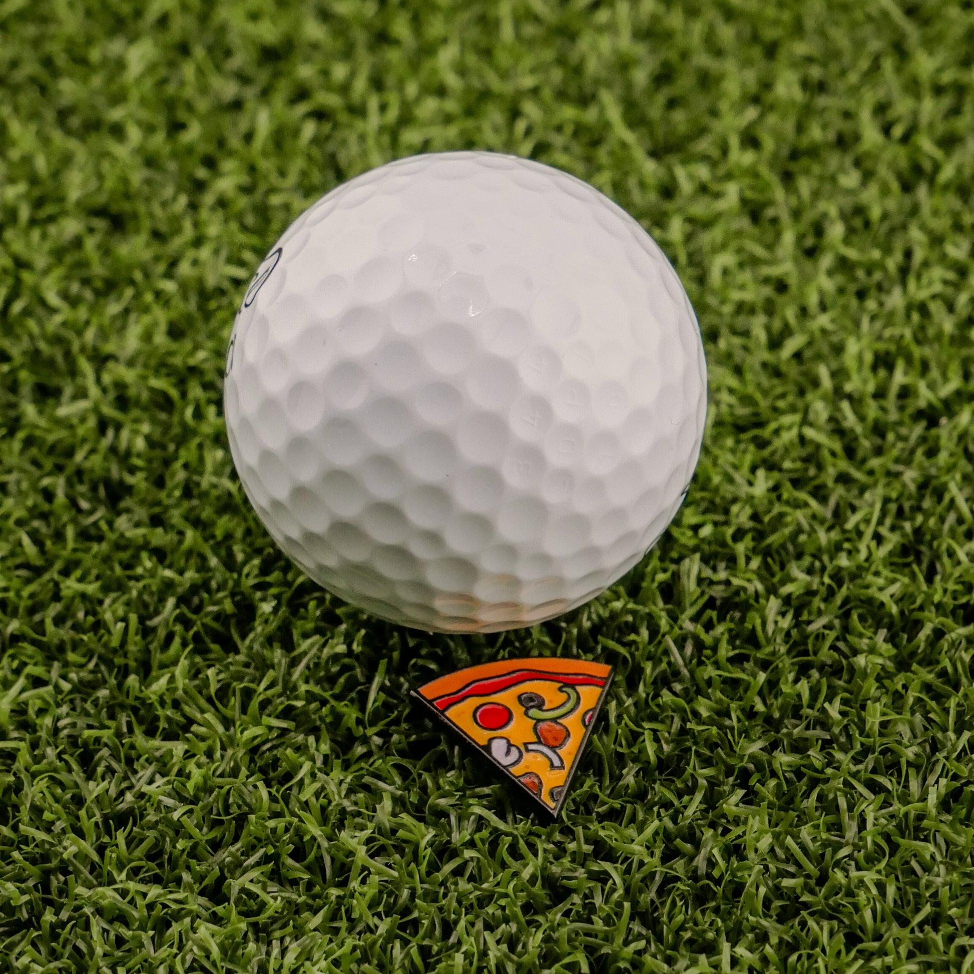 A white golf ball sits on green turf beside the Logie Joe's Golf Supreme Pizza "Slice" Premium Ball Marker, designed as a small triangular pizza slice with pepperoni and cheese.