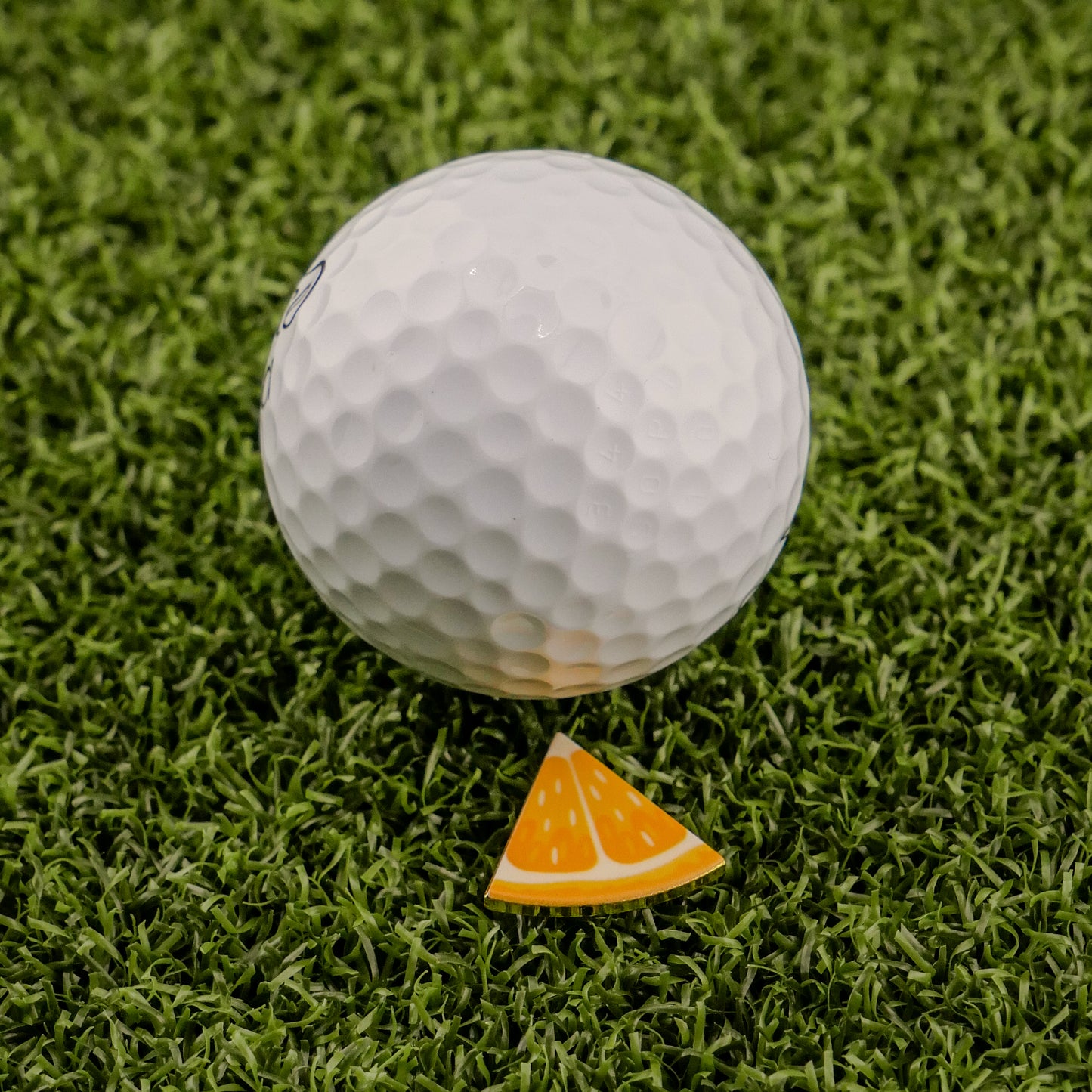 A white golf ball is on green artificial grass beside the Logie Joe's Golf Orange "Slice" Premium Ball Marker, a small, triangular orange marker designed to look like a removable slice.