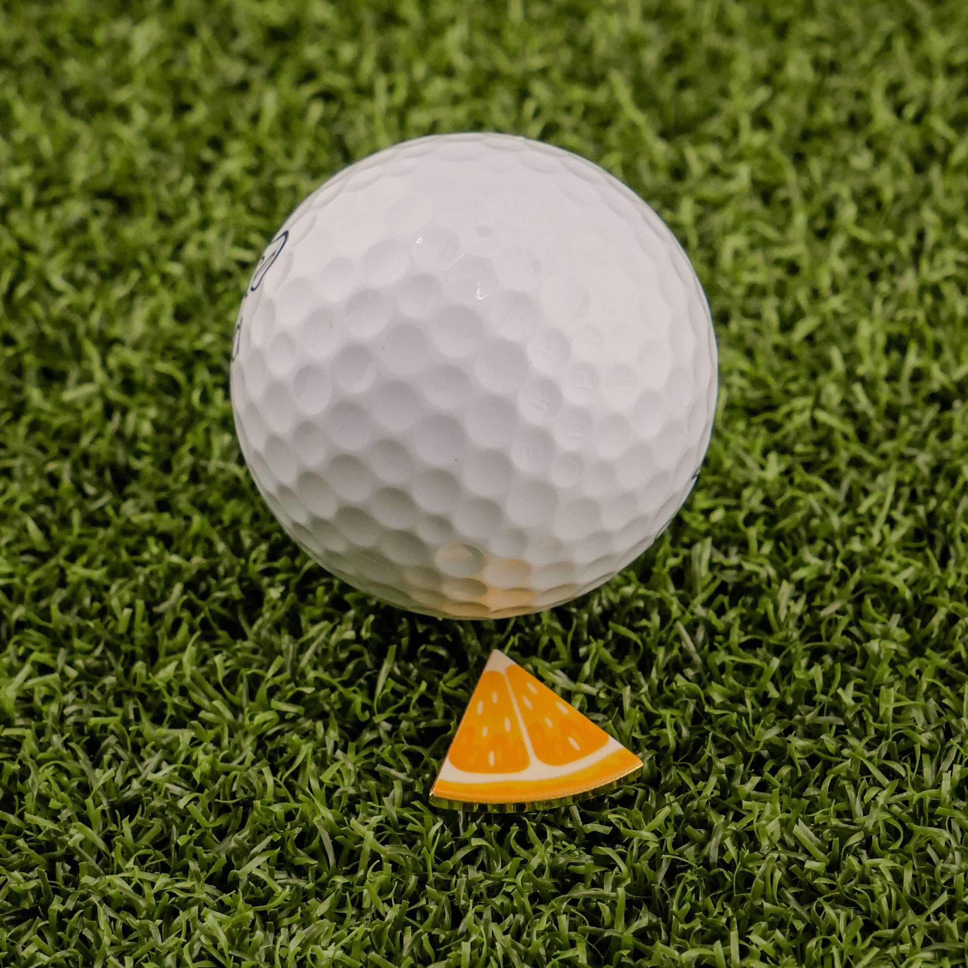 A white golf ball is on green artificial grass beside the Logie Joe's Golf Orange "Slice" Premium Ball Marker, a small, triangular orange marker designed to look like a removable slice.