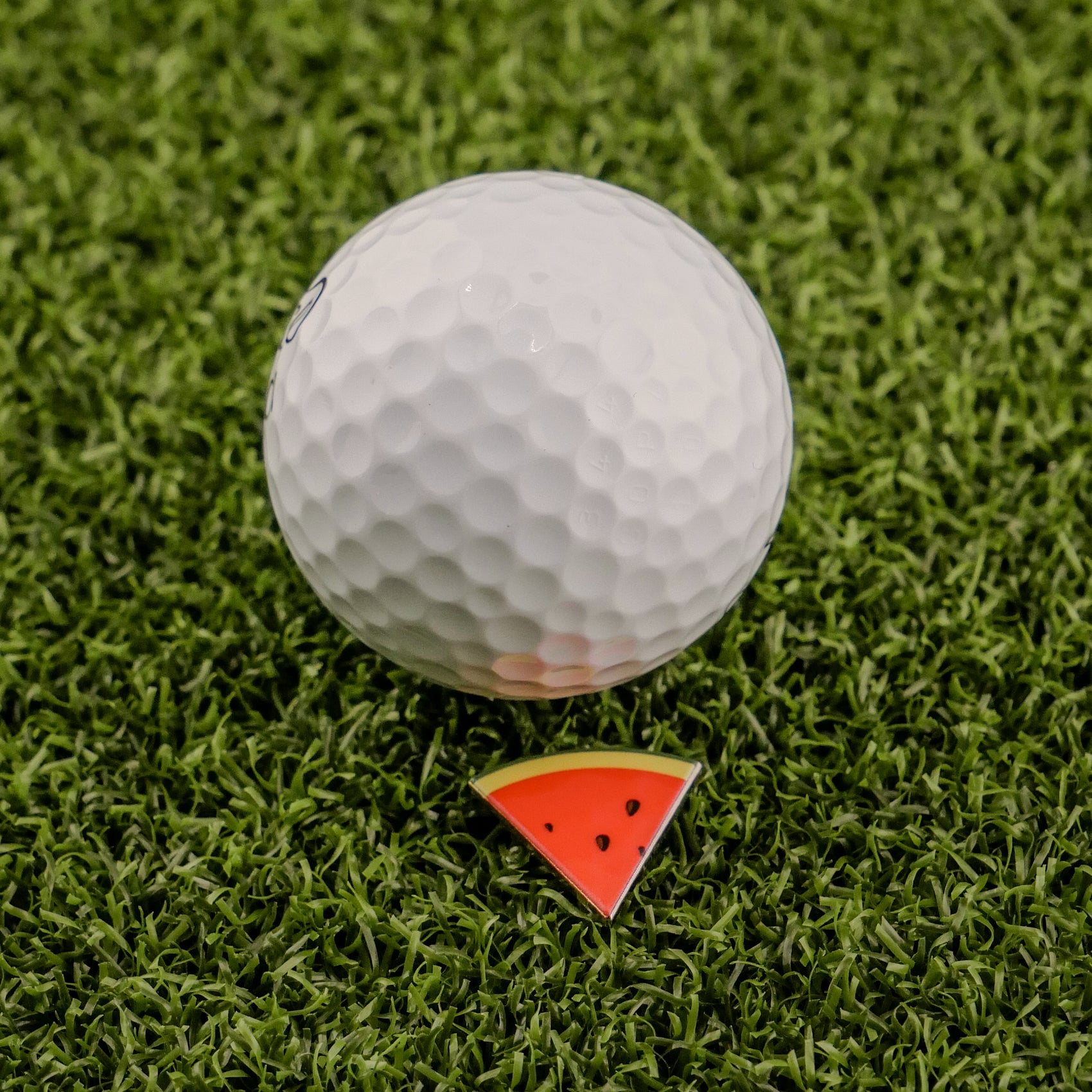 A white golf ball rests on artificial green grass beside Logie Joe's Golf Watermelon "Slice" Premium Ball Marker, a small triangular marker resembling a watermelon slice.