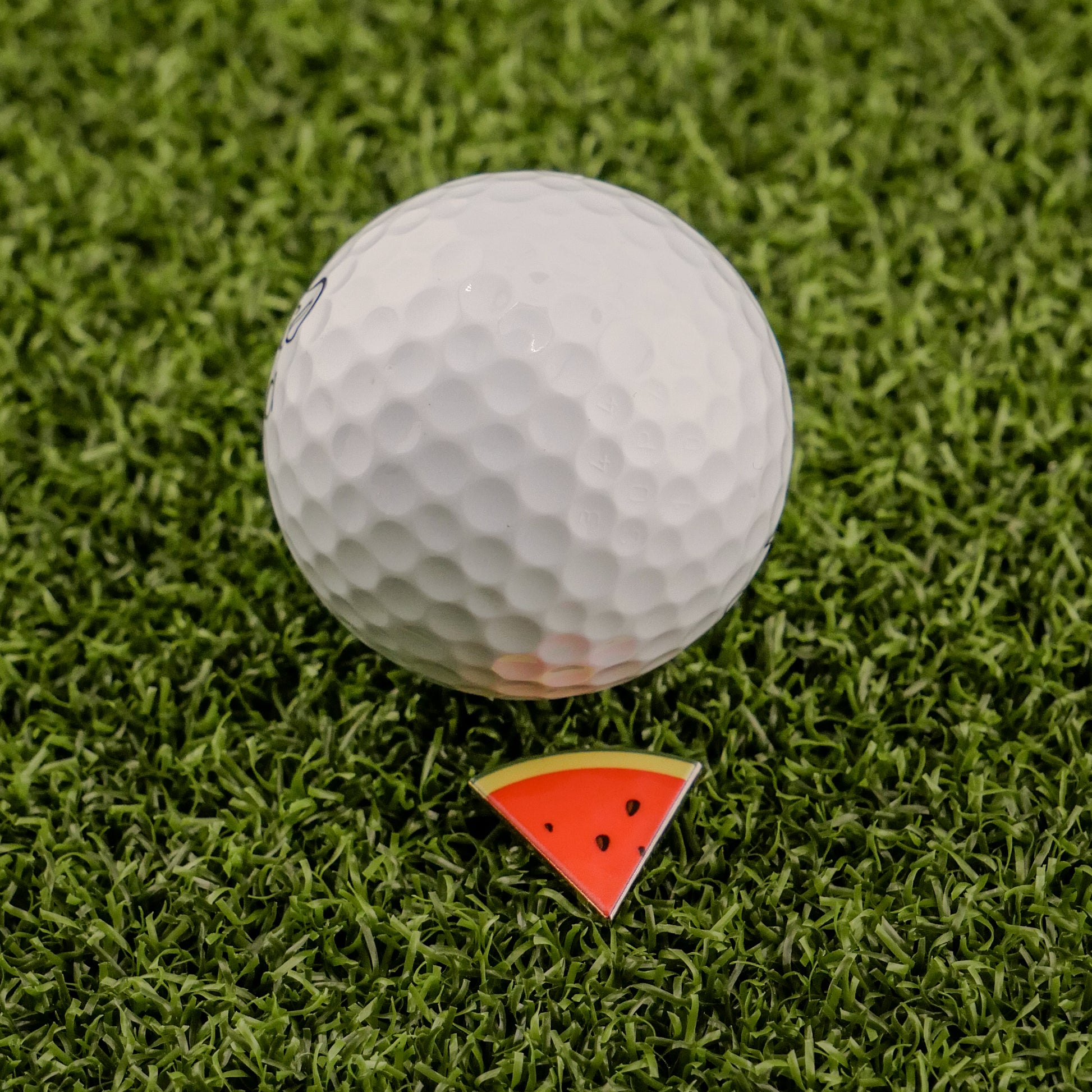 A white golf ball rests on artificial green grass beside Logie Joe's Golf Watermelon "Slice" Premium Ball Marker, a small triangular marker resembling a watermelon slice.