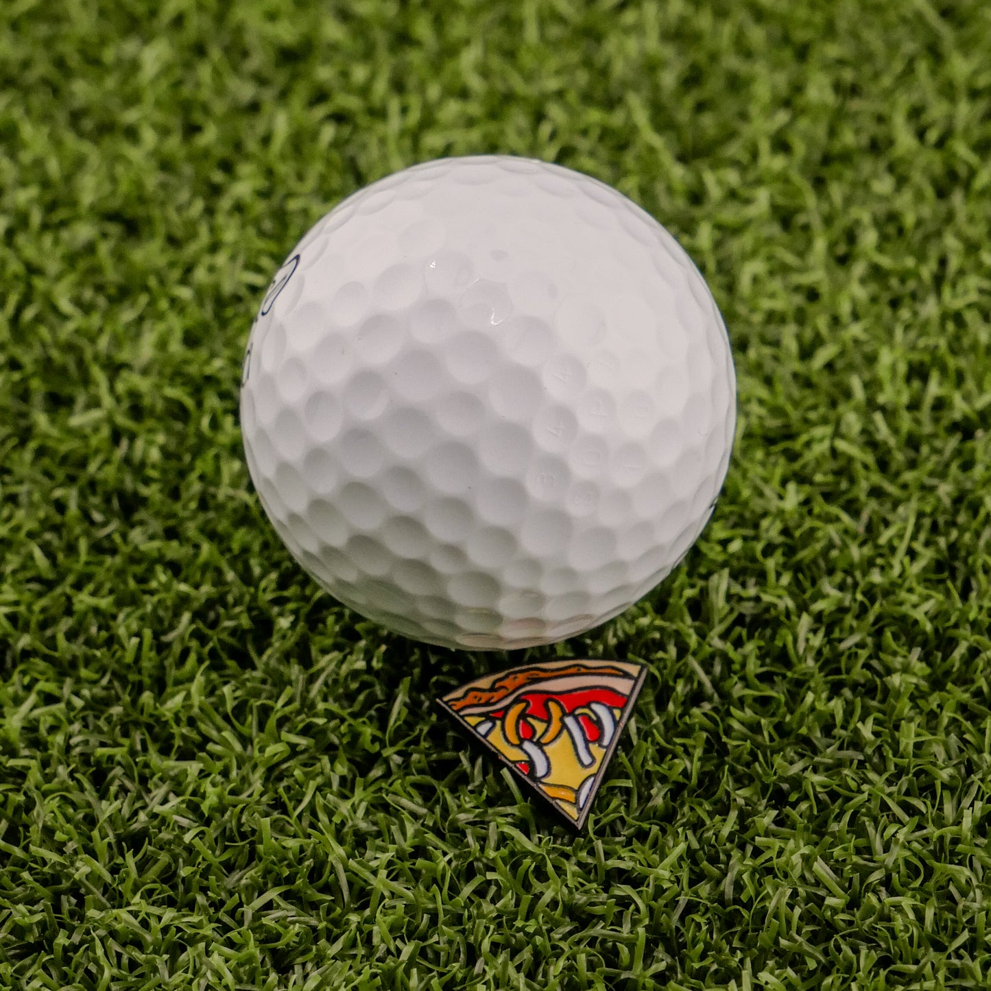 A white golf ball sits on Logie Joe's Golf Mexican Pizza "Slice" Premium Ball Marker, which displays a vibrant design atop bright green artificial turf.