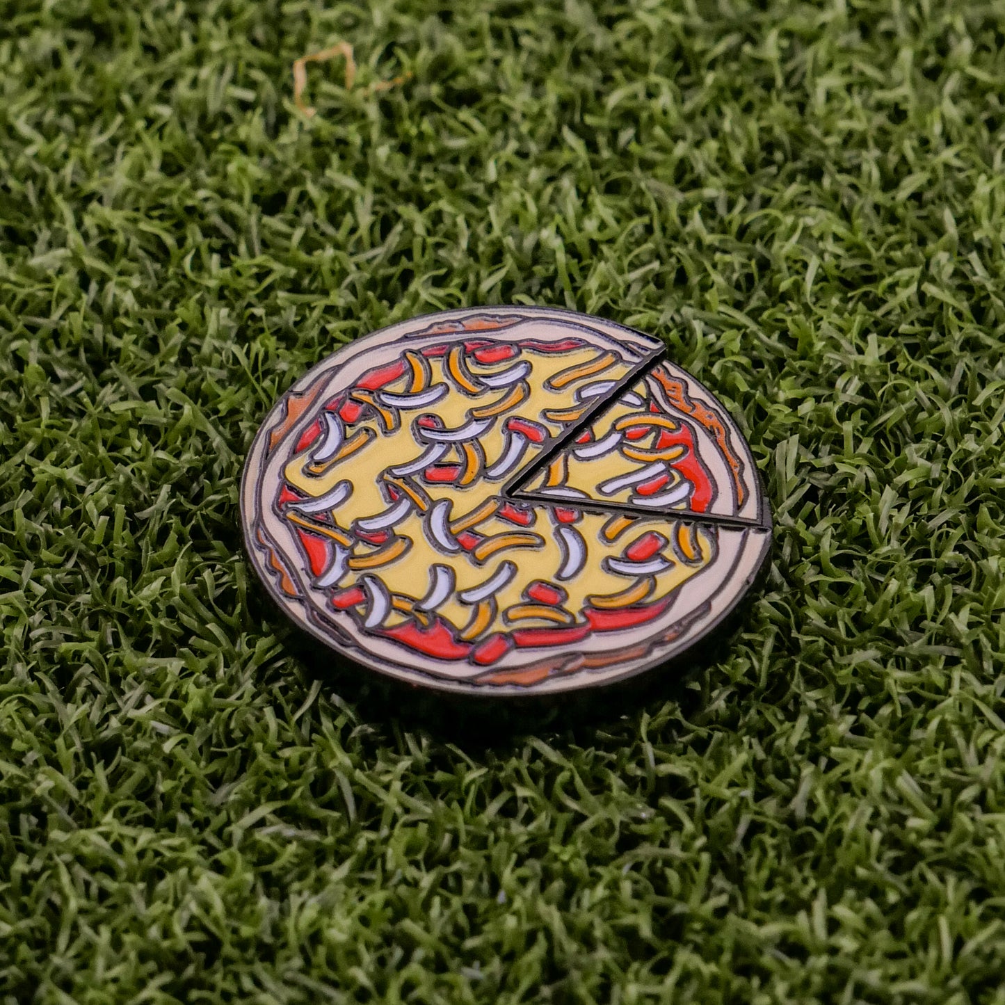 The Logie Joe's Golf Mexican Pizza "Slice" Premium Ball Marker is a round, cartoon-style pin with a removable slice, featuring yellow cheese and red-white toppings, shown on green artificial grass for a playful touch.
