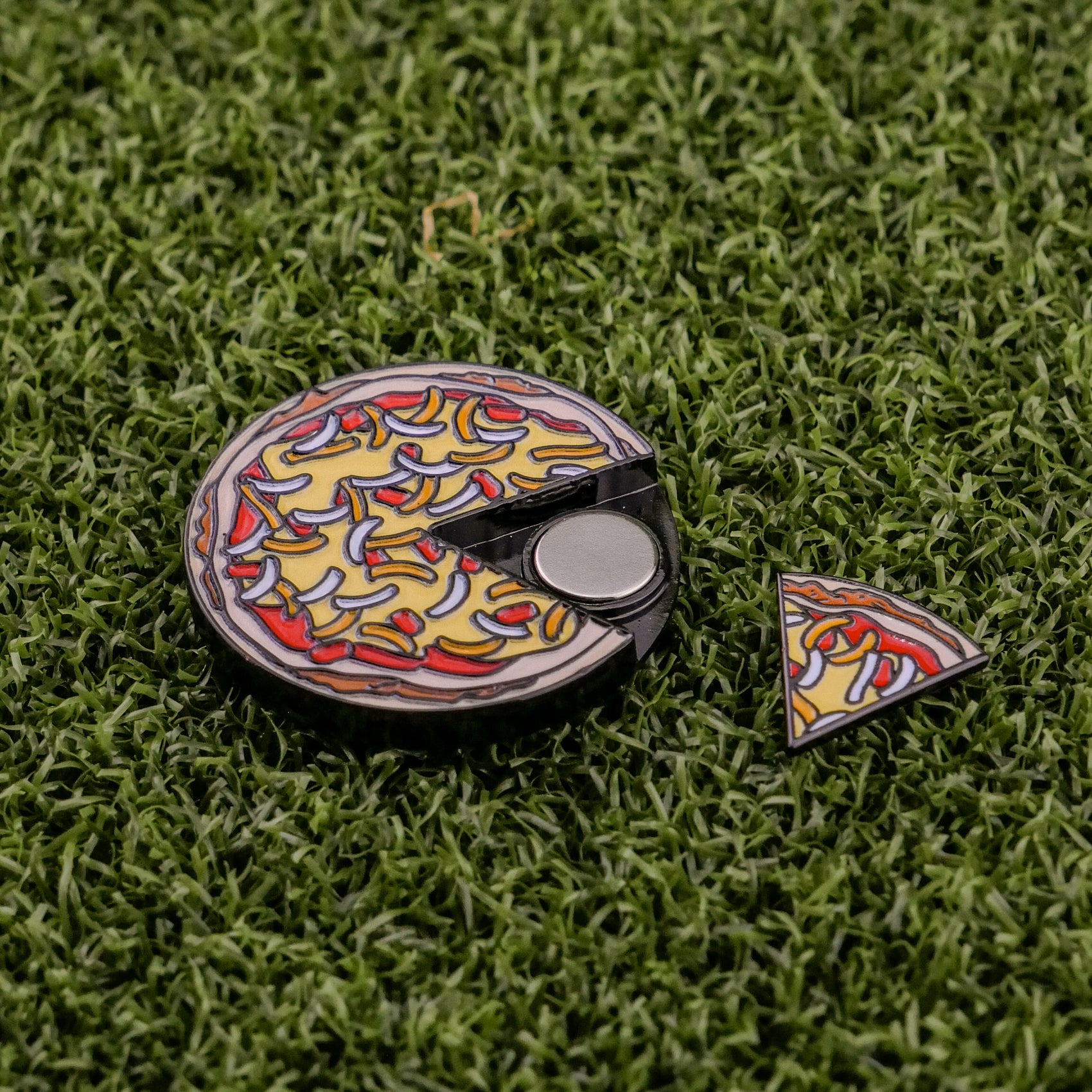 The Logie Joe's Golf Mexican Pizza "Slice" Premium Ball Marker, featuring a removable slice and vibrant toppings, adds a playful touch as it rests on green artificial grass.