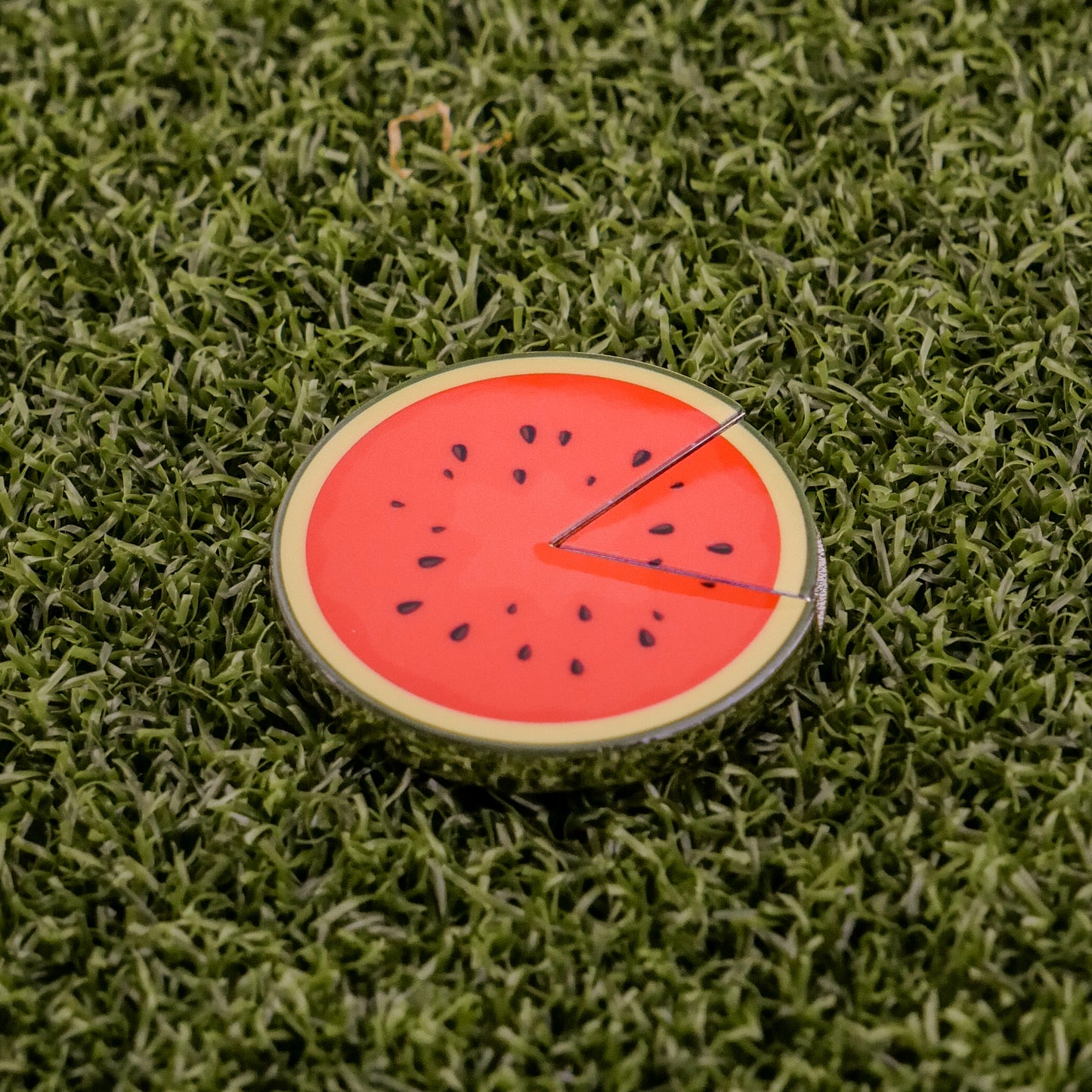 Logie Joe's Golf Watermelon "Slice" Premium Ball Marker features a red center, black seeds, green rind, and removable slice. This fun accessory resembles a watermelon on green turf—perfect for adding personality to your golf game.