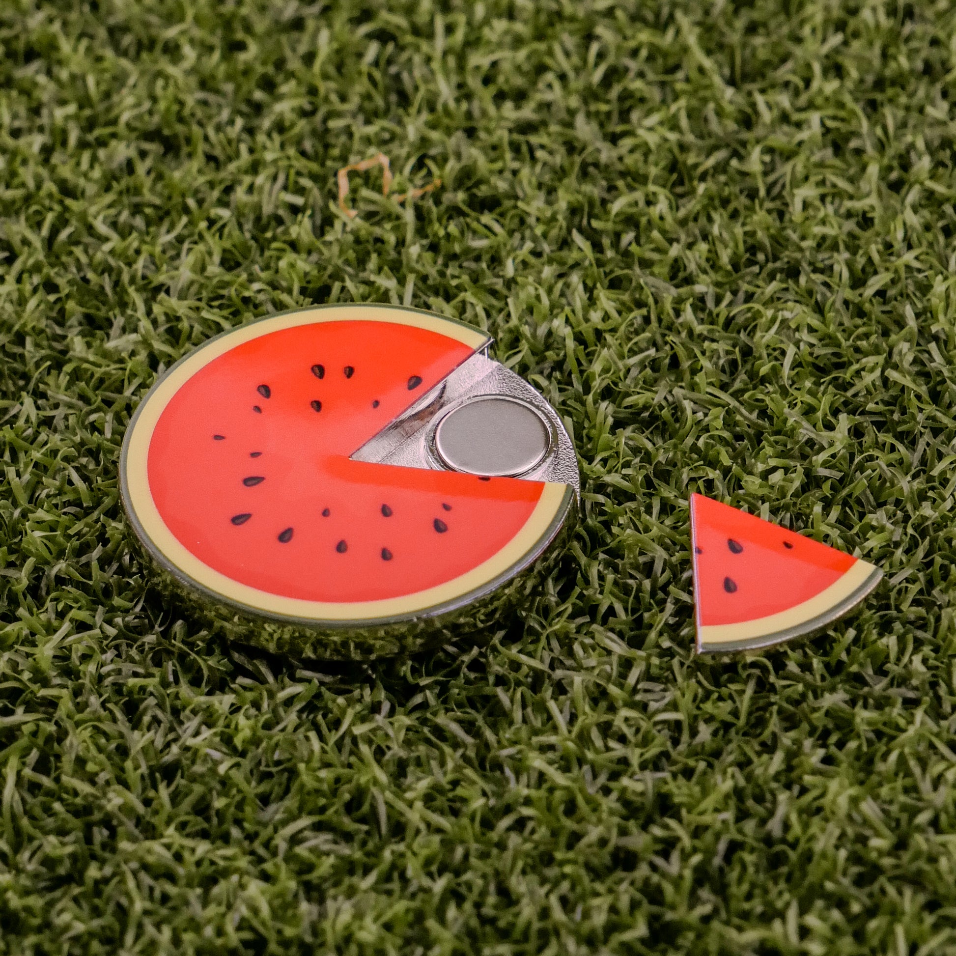 The Logie Joe's Golf Watermelon "Slice" Premium Ball Marker, shaped like a watermelon slice with a removable section revealing a silver coin, rests on green artificial grass with the removed piece beside it.