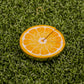 The Orange "Slice" Premium Ball Marker by Logie Joe's Golf, shaped like an orange slice with two small cracks, rests on green artificial grass as a distinctive golf ball marker.