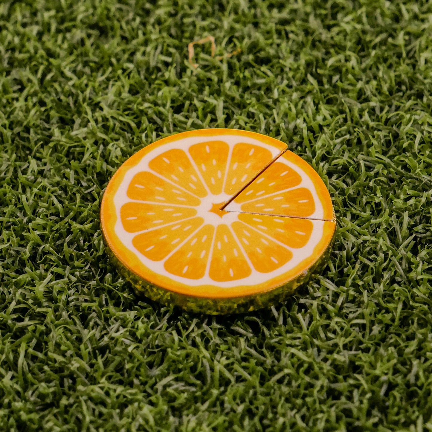 The Orange "Slice" Premium Ball Marker by Logie Joe's Golf, shaped like an orange slice with two small cracks, rests on green artificial grass as a distinctive golf ball marker.