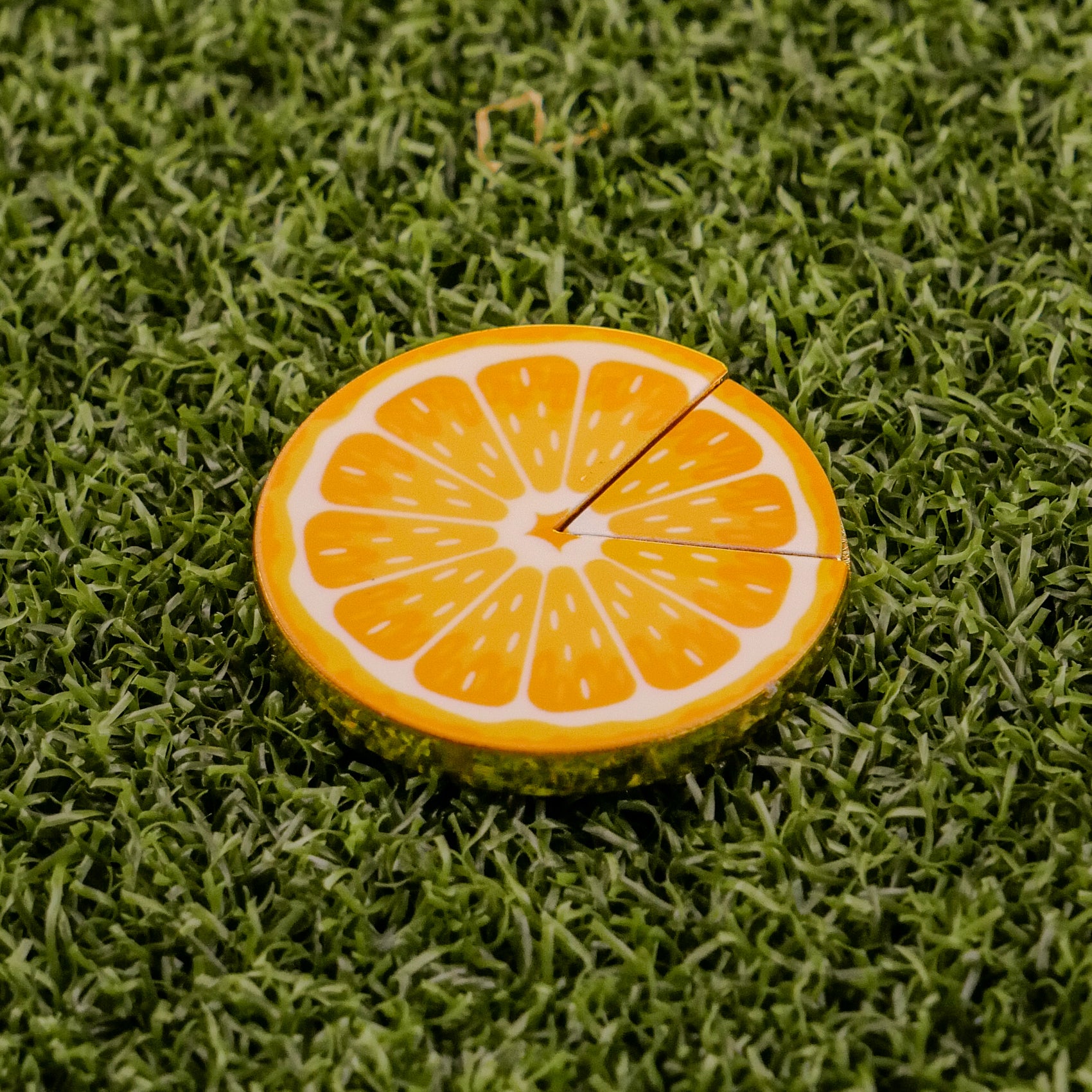 The Orange "Slice" Premium Ball Marker by Logie Joe's Golf, shaped like an orange slice with two small cracks, rests on green artificial grass as a distinctive golf ball marker.