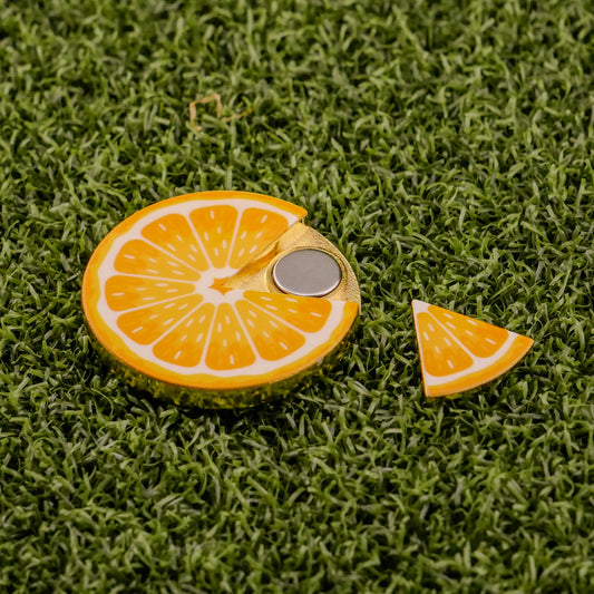 The Logie Joe's Golf Orange "Slice" Premium Ball Marker features a round orange slice design with a metallic center and removable piece, shown on green artificial grass.