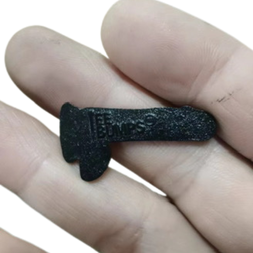 A hand holds a small, black, T-shaped plastic ball marker from TeeBumps called The Dildy Ball Marker, embossed with THE BUMPS.