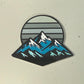 The Mountain Moonrise Ball Marker from Logie Joe's Golf features a stylized illustration of snow-capped mountains with blue accents set against a gray sun with horizontal lines on a light background, creating a unique and nature-inspired golf marker that’s perfect for outdoor golf enthusiasts.