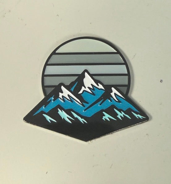 The Mountain Moonrise Ball Marker from Logie Joe's Golf features a stylized illustration of snow-capped mountains with blue accents set against a gray sun with horizontal lines on a light background, creating a unique and nature-inspired golf marker that’s perfect for outdoor golf enthusiasts.