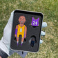 A hand in a white golf glove holds the Pin Creatures MAMBA Divot Tool tin, featuring cartoon #8 yellow and #24 purple jersey pins plus a ball marker set in black foam, with grass visible in the background.