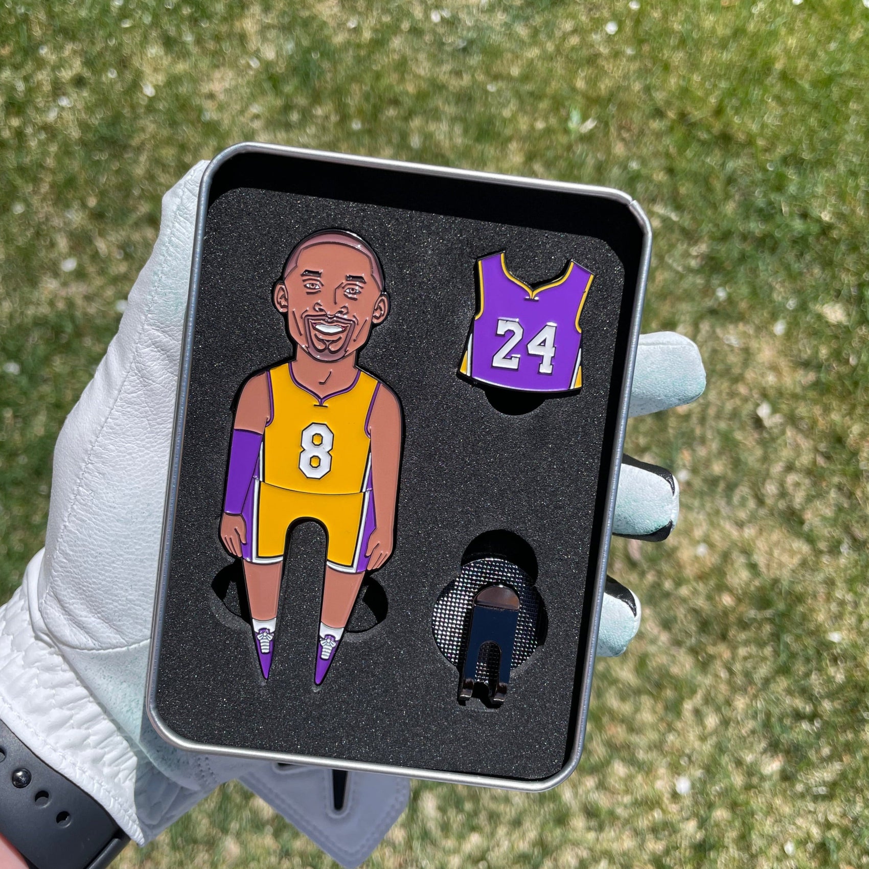 A hand in a white golf glove holds the Pin Creatures MAMBA Divot Tool tin, featuring cartoon #8 yellow and #24 purple jersey pins plus a ball marker set in black foam, with grass visible in the background.