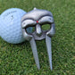 A Guerrilla Golf Gladiator Divot Tool, shaped like a stylized mask and finished in silver, rests on the grass beside a white Titleist 3 golf ball.
