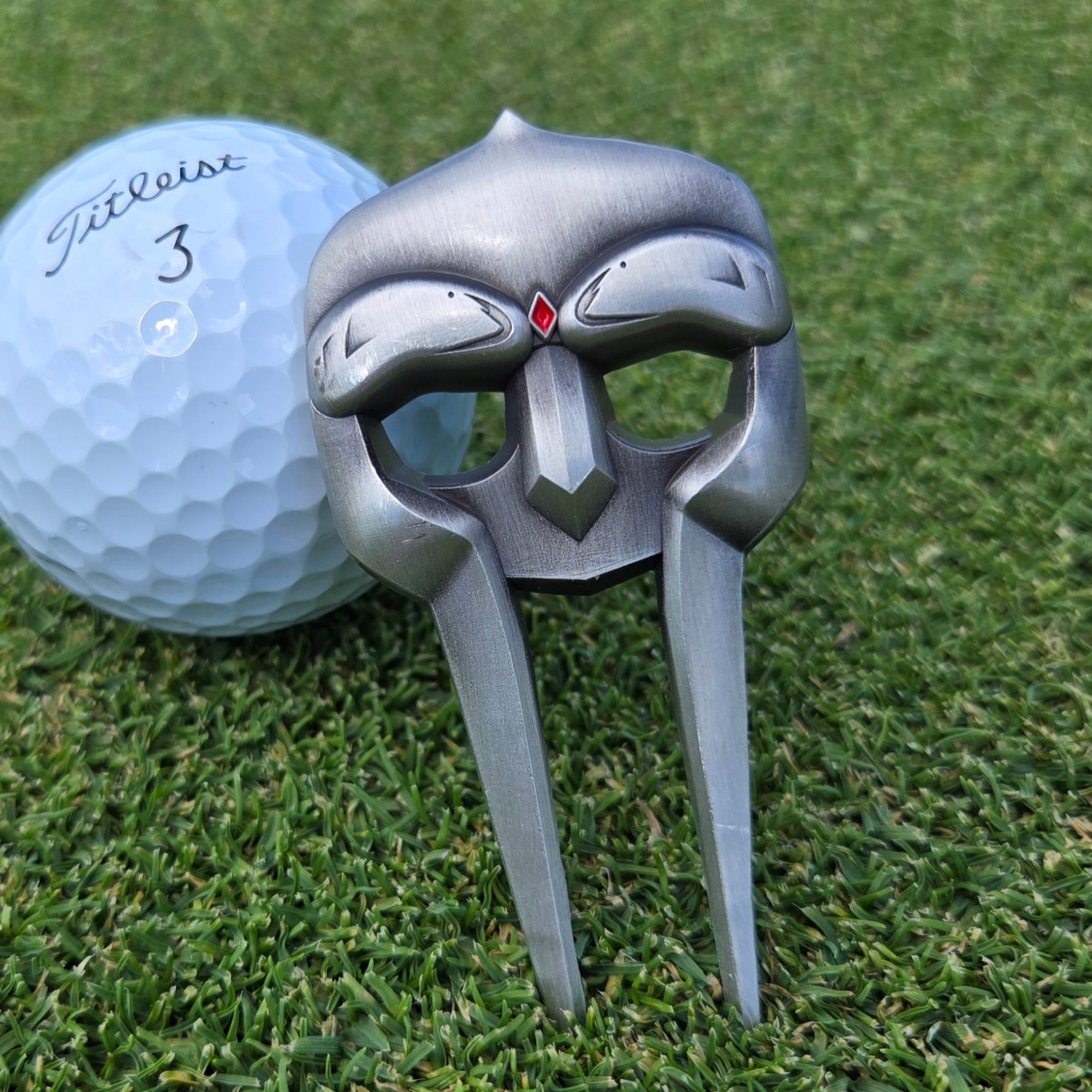 A Guerrilla Golf Gladiator Divot Tool, shaped like a stylized mask and finished in silver, rests on the grass beside a white Titleist 3 golf ball.
