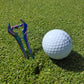 The Guerrilla Golf E Sword Divot Tool, with its shiny iridescent finish, stands upright beside a white golf ball on neatly trimmed green grass—making it an essential accessory for any golfer.