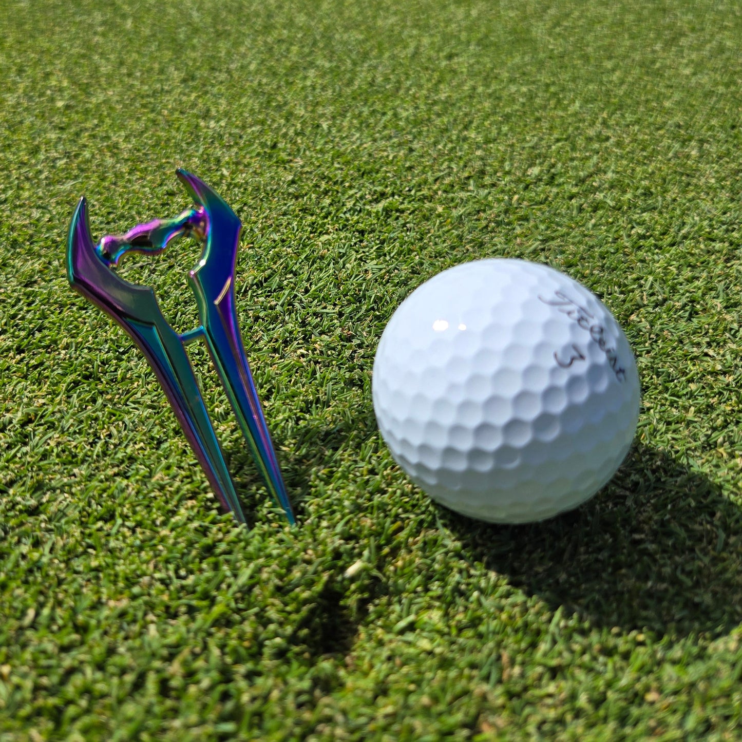 The Guerrilla Golf E Sword Divot Tool, with its shiny iridescent finish, stands upright beside a white golf ball on neatly trimmed green grass—making it an essential accessory for any golfer.