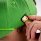 A person attaches the Pins and Aces Pimento Cheese Sandwich Ball Marker to the back of a bright green cap.