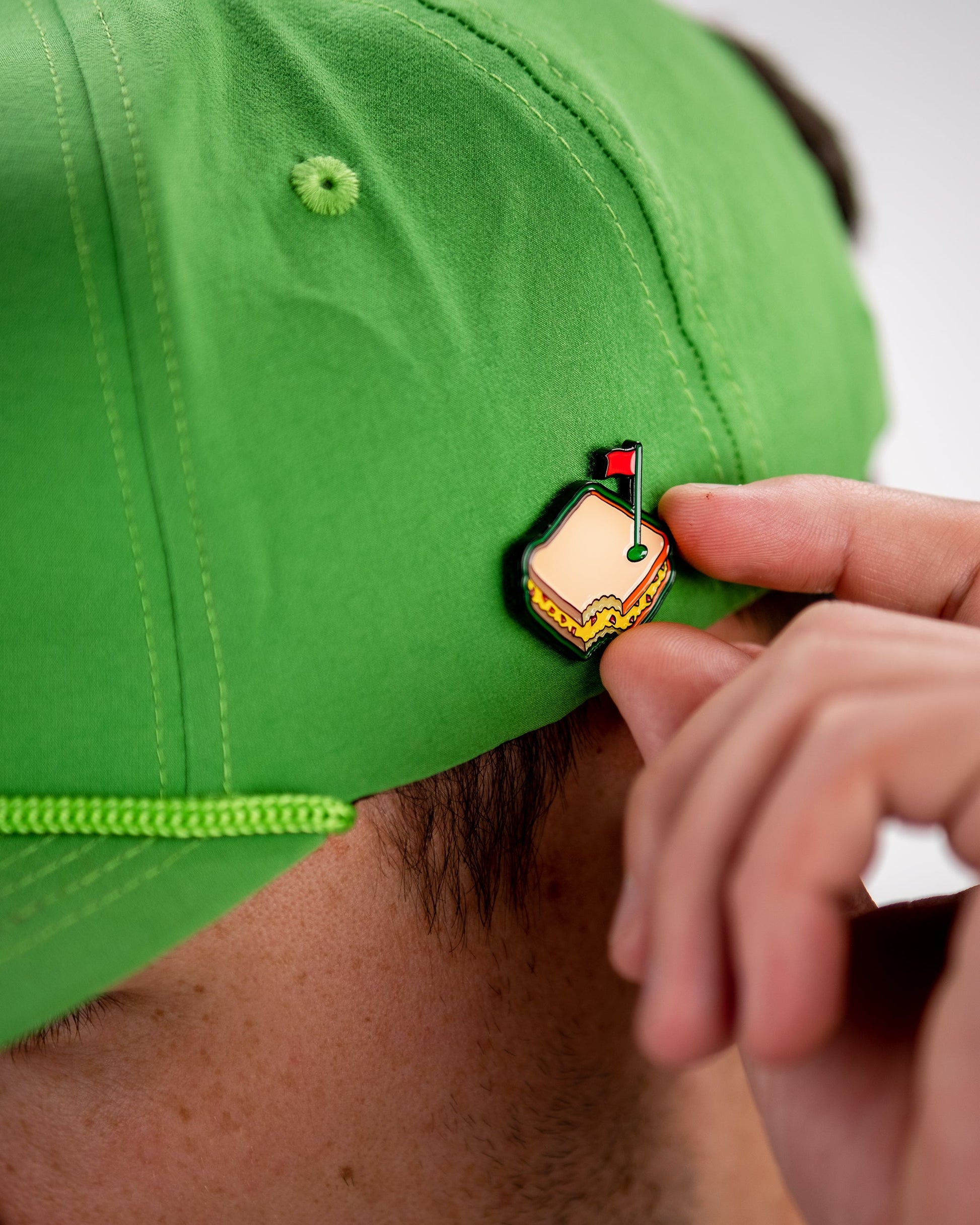 A person attaches the Pins and Aces Pimento Cheese Sandwich Ball Marker to the back of a bright green cap.