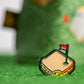 The Pins and Aces Pimento Cheese Sandwich Ball Marker, shaped like a sandwich with a bite and topped with a red golf flag, rests on green turf—a fun Augusta-inspired golf accessory.