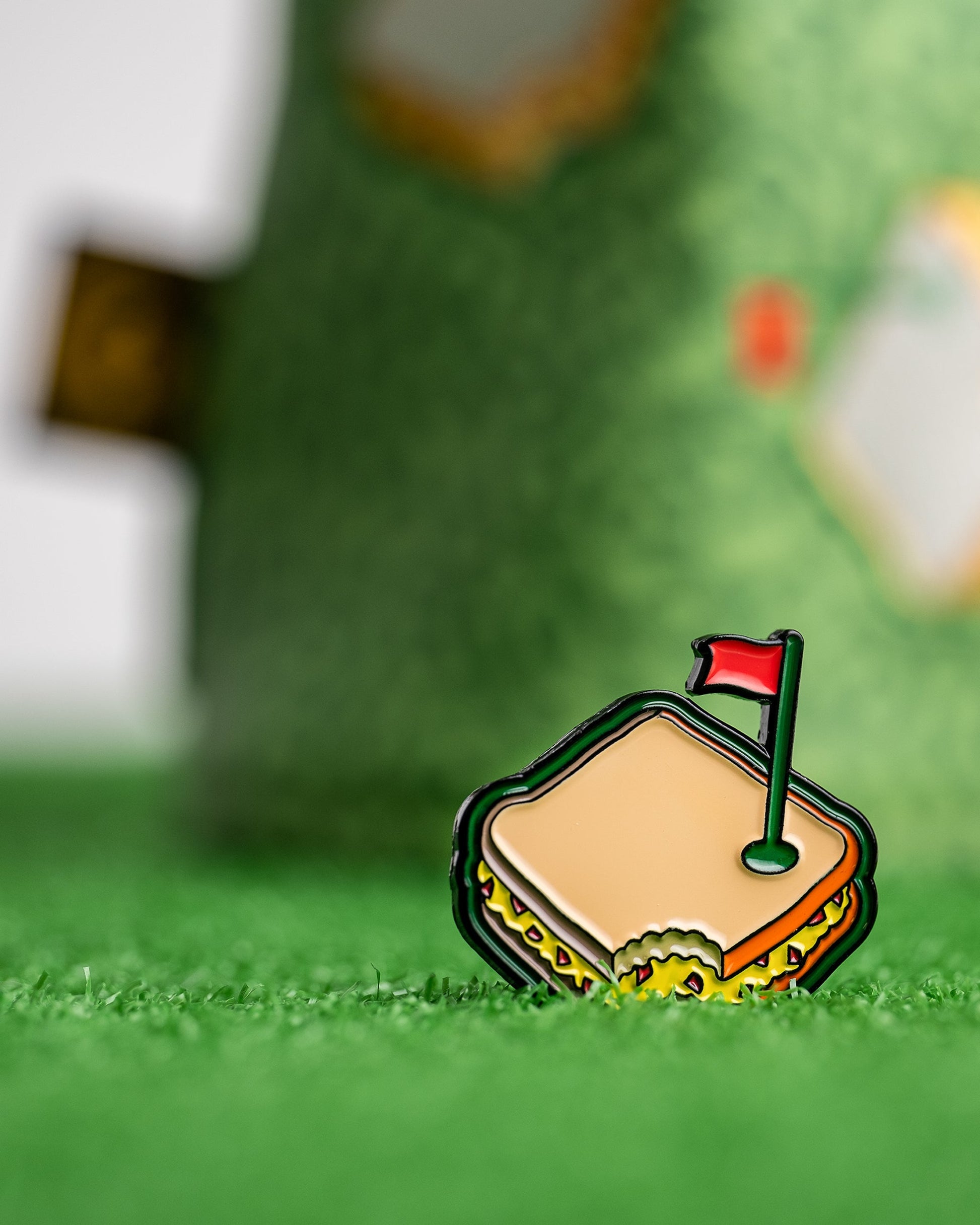 The Pins and Aces Pimento Cheese Sandwich Ball Marker, shaped like a sandwich with a bite and topped with a red golf flag, rests on green turf—a fun Augusta-inspired golf accessory.