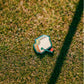 A Pimento Cheese Sandwich Ball Marker by Pins and Aces, styled like an Augusta sign with a red and black handle, rests on green grass in sunlight, casting a shadow.