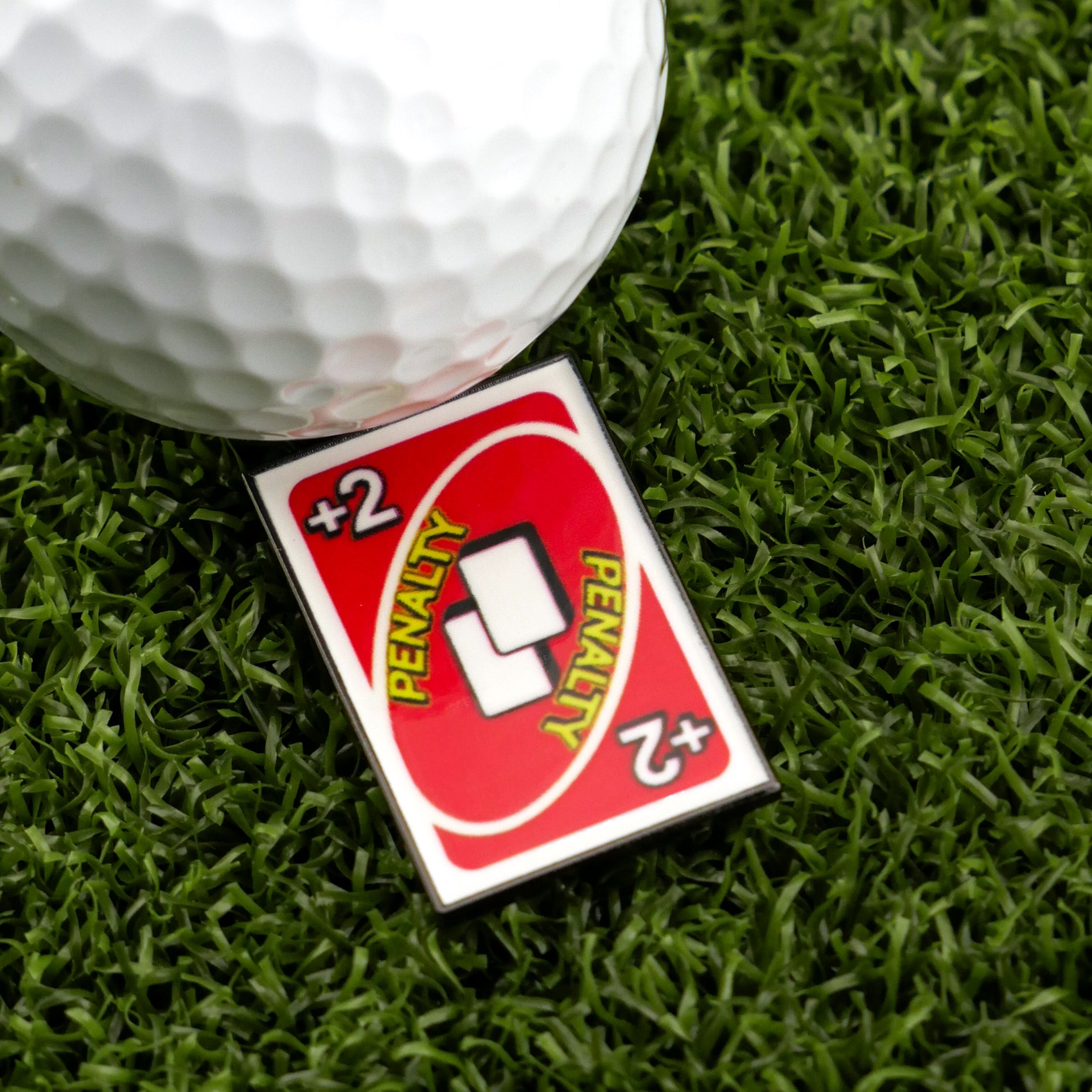 A close-up of a golf ball on artificial grass next to Logie Joe's Golf 2 Penalty Card - Red, one of the most humorous and unique premium golf ball markers you’ll find.