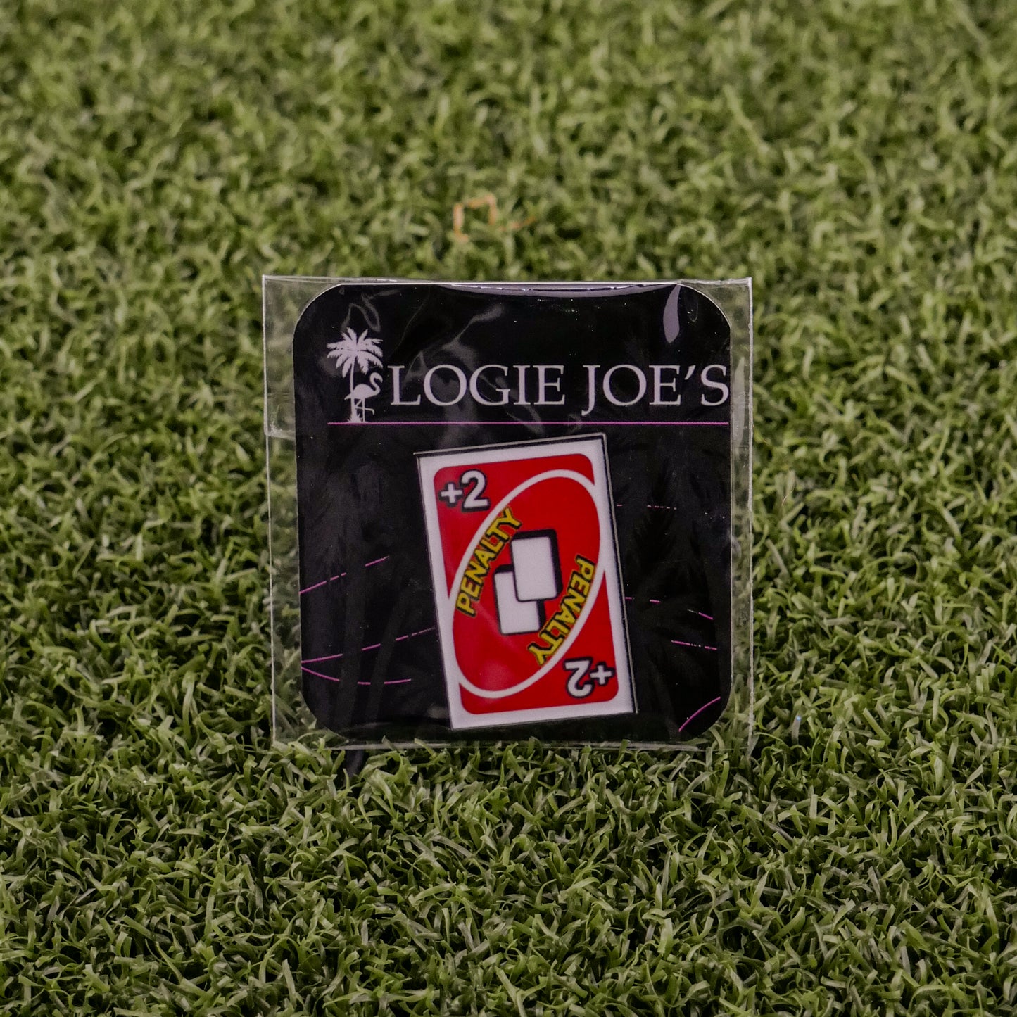 The 2 Penalty Card - Red enamel pin by Logie Joe's Golf displays a red "+2 PENALTY" Uno card on a branded backing, set on green turf—ideal for golf fans seeking a fun, quirky alternative to standard ball markers.
