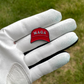 A white golf glove holds a small red Pin Creatures MAGA Hat Ball Marker shaped like a baseball cap with "MAGA" in white letters; blurred grass can be seen in the background.