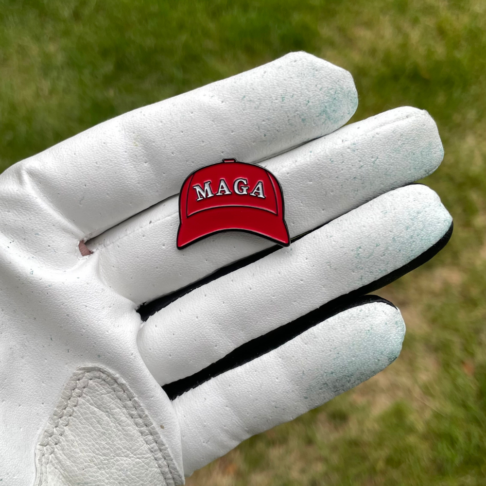 A white golf glove holds a small red Pin Creatures MAGA Hat Ball Marker shaped like a baseball cap with "MAGA" in white letters; blurred grass can be seen in the background.