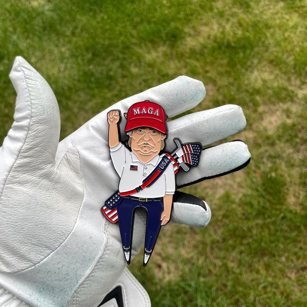 A gloved hand holds the Pin Creatures MAGA Hat Ball Marker, featuring a cartoon man in a red MAGA hat, white shirt, and blue pants raising his fist with an American flag golf club against a grassy background.