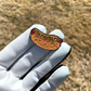 A gloved hand holds the Pin Creatures Glizzy Ball Marker, shaped like a hot dog with "Glizzy" in yellow script on the bun, against a blurred brown grass background.