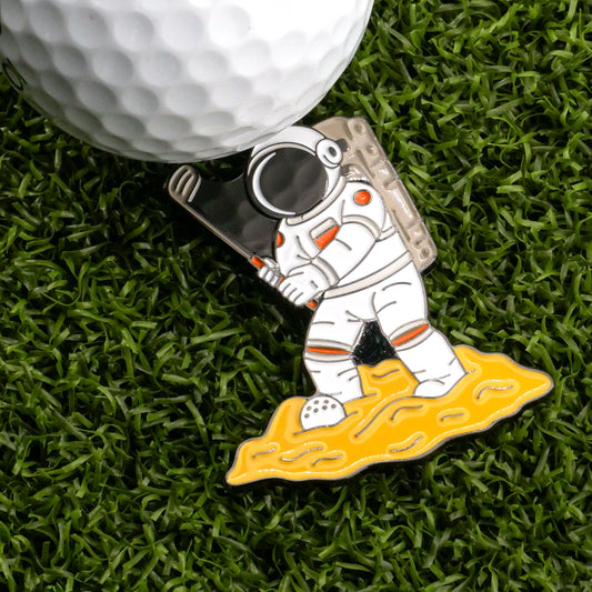 Ballmarkers.com’s Giant Swing for Mankind Ball Marker features an astronaut holding a golf club on a yellow, cheese-like surface—ideal for marking your spot on artificial green grass next to a white golf ball.
