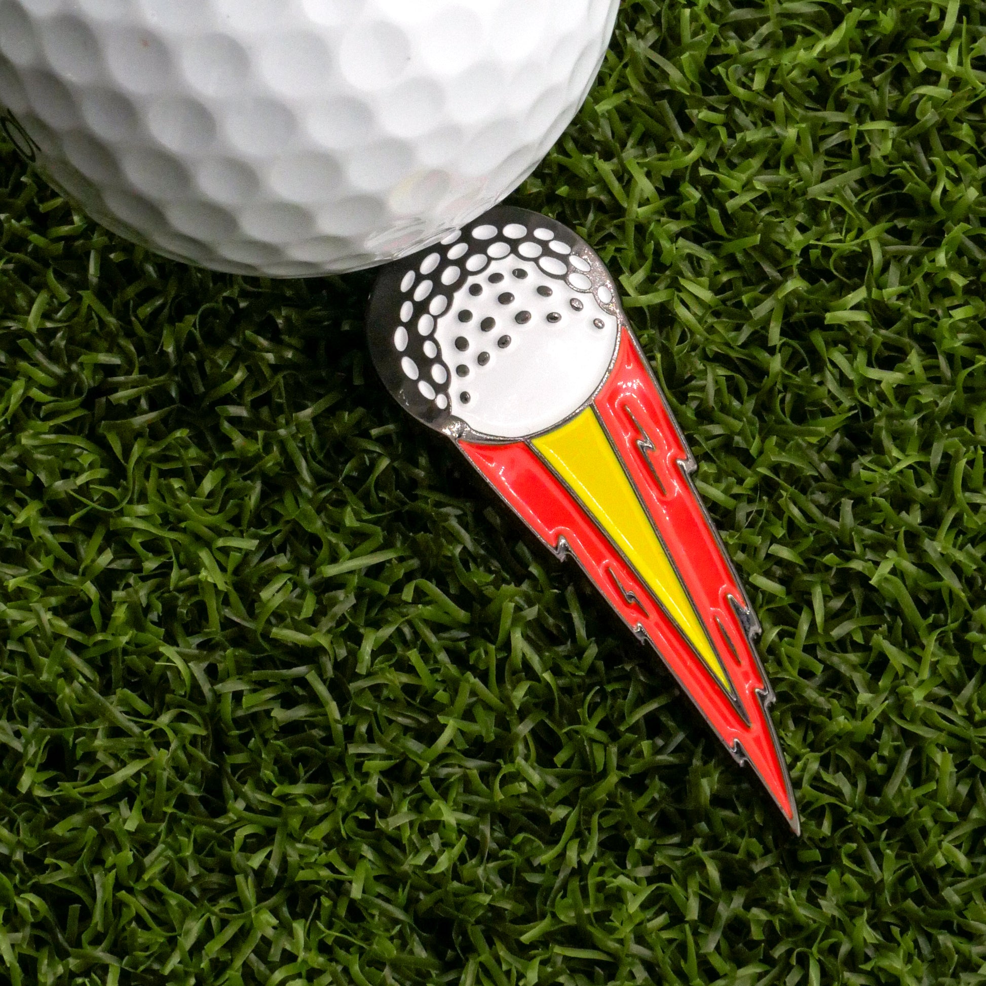 A close-up of a golf ball on artificial grass next to the Ballmarkers.com Hot Shot Tracer Ball Marker, a red, yellow, and silver marker shaped like a golf ball with a fiery tail—ideal for marking your spot after a long drive.