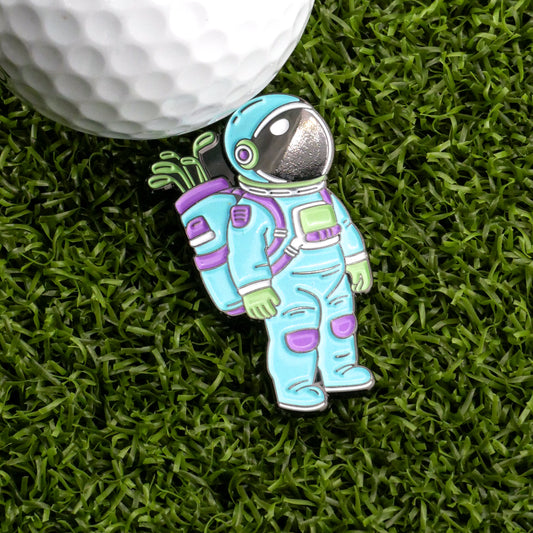 The Astronaut Space Walker Ball Marker from Ballmarkers.com features a colorful astronaut with golf clubs in a backpack, shown on green artificial grass beside a white golf ball—ideal for fans of Ballmarkers.com.